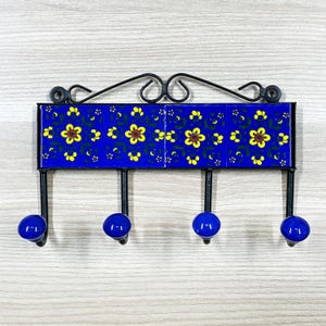 Könnte beinhalten: Ein an der Wand montierter Schlüsselhalter mit vier Haken. Der Halter hat ein blaues Keramikfliesen-Design mit gelben Blumenmustern. Die Haken sind mit blauen Knöpfen versehen und an einem schwarzen Metallrahmen mit dekorativen Wirbeln befestigt.