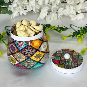 Op de afbeelding: Een kleurrijke keramische pot met een deksel, gevuld met cashewnoten. De pot heeft een geometrisch patroon met verschillende kleuren, waaronder blauw, groen, geel en rood.