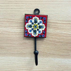 May include: A black metal hook with a red and blue ceramic tile featuring a floral design. The tile has a white centre with yellow flowers and blue and green leaves.