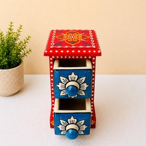 Può includere: Una piccola cassettiera in legno con una struttura rosso vivace e dettagli a pois bianchi. Include due cassetti estraibili con frontali blu brillante, ciascuno decorato con un motivo floreale bianco e un pomello circolare blu tinta unita.