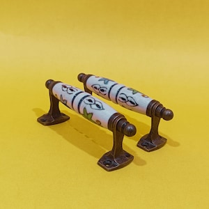 Könnte beinhalten: Zwei Keramik-Türgriffe mit floralem Design. Die Griffe sind weiß mit braunen und grünen Akzenten. Die Griffe sind an Messingplatten befestigt.