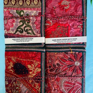 Puede incluir: Cuatro cuadernos hechos a mano con coloridas cubiertas de tela bordada. Las cubiertas presentan tonos rojos, dorados y verdes con patrones intrincados. Cada cuaderno está atado con un cordón decorativo e incluye una etiqueta que dice "HAND MADE PAPER NOTE BOOK".