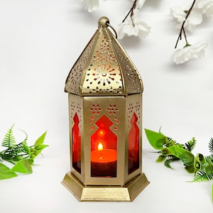 Può includere: Una lanterna in metallo dorato con intricati disegni ritagliati e un inserto in vetro rosso. Una piccola candela brucia all'interno della lanterna.