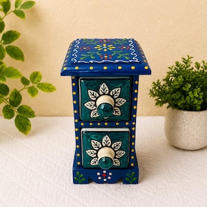 May include: A small blue wooden chest with two drawers featuring teal ceramic fronts. Each drawer has a white floral pattern and a round knob. The blue frame is decorated with yellow dots and green leaf motifs, with a floral design on the top surface.