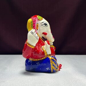 May include: A colourful, hand-painted metal figurine of Ganesha, the Hindu god of wisdom and prosperity. The figurine is sitting with its trunk raised and its hands in a gesture of blessing. It is painted in shades of red, blue, white, and gold.