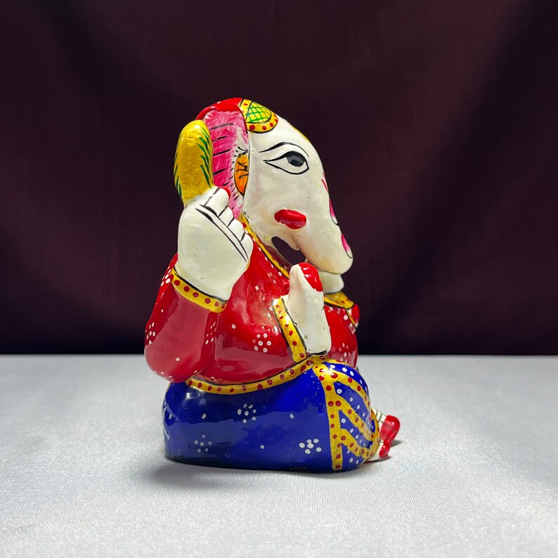May include: A colourful, hand-painted metal figurine of Ganesha, the Hindu god of wisdom and prosperity. The figurine is sitting with its trunk raised and its hands in a gesture of blessing. It is painted in shades of red, blue, white, and gold.