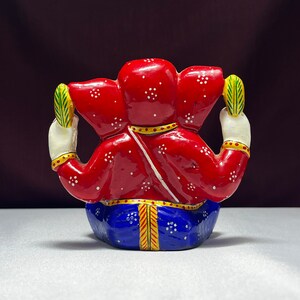 May include: A red and blue painted figurine of the Hindu deity Ganesha. The figurine is sitting with its legs crossed and its hands raised in a gesture of blessing. The figurine is decorated with white dots and gold accents.