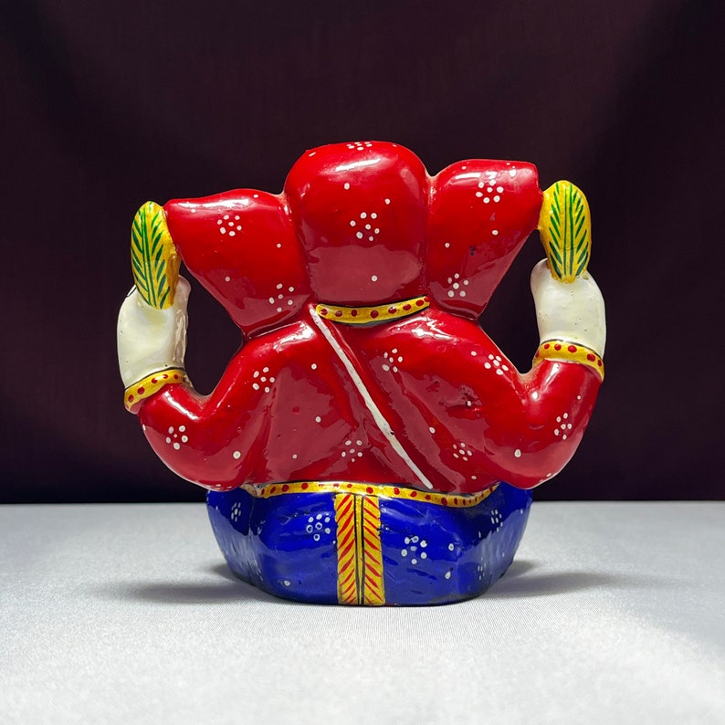 May include: A red and blue painted figurine of the Hindu deity Ganesha. The figurine is sitting with its legs crossed and its hands raised in a gesture of blessing. The figurine is decorated with white dots and gold accents.