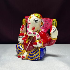May include: A colourful, hand-painted metal figurine of Ganesha, the Hindu god of wisdom and prosperity. The figurine is sitting with its legs crossed, and its trunk is curled up. The figurine is decorated with intricate details, including flowers, jewels, and a red and blue patterned robe.