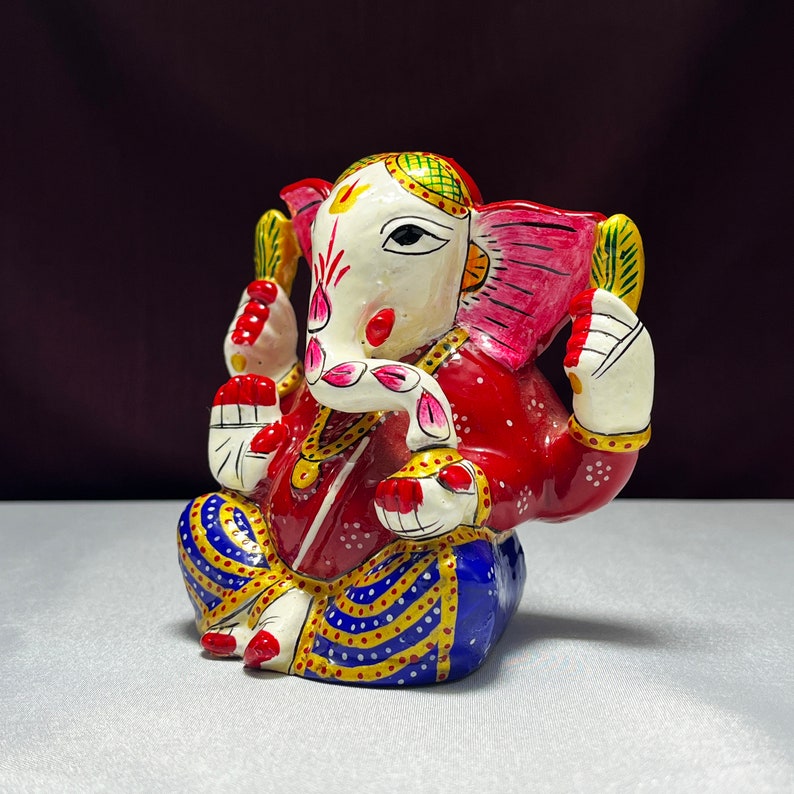 May include: A colourful, hand-painted metal figurine of Ganesha, the Hindu god of wisdom and prosperity. The figurine is sitting with its legs crossed, and its trunk is curled up. The figurine is decorated with intricate details, including flowers, jewels, and a red and blue patterned robe.