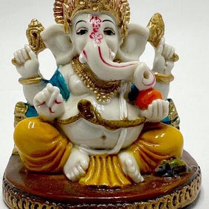 Op de afbeelding: Gedetailleerd beeldje van de hindoeïstische godheid Ganesha, zittend met meerdere armen. Het beeld heeft een wit lichaam, gouden accenten en een rode en gouden kroon. Ganesha houdt een kleine oranje vrucht vast. De basis is bruin met gouden randen.