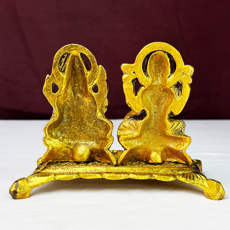 May include: Gold-colored metal statue of a Hindu deity couple, Lakshmi and Ganesha, sitting on a decorative base. The statue is intricately detailed and has a traditional design.