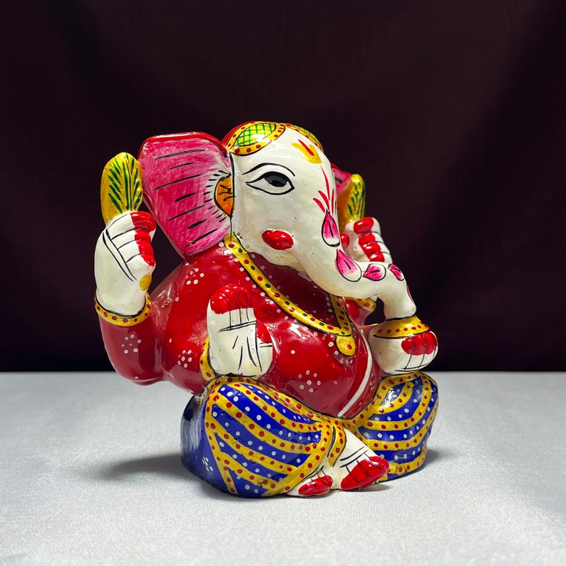 May include: A colourful hand-painted metal figurine of the Hindu deity Ganesha. The figurine is red, white, blue, yellow, and green. Ganesha is depicted sitting with his trunk raised and his hands in a mudra.