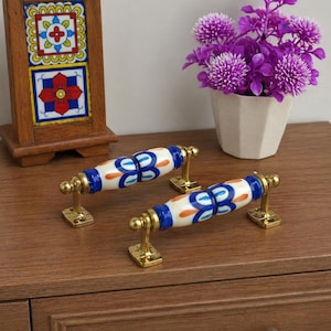 Peut inclure: Deux poignées de meubles décoratives avec un motif floral blanc, bleu et orange. Les poignées ont une quincaillerie de montage de couleur laiton. Les poignées sont sur une surface en bois.