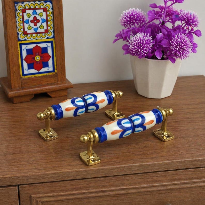 May include: Two decorative cabinet handles with a white, blue, and orange floral design. The handles have brass-coloured mounting hardware. The handles are on a wooden surface.