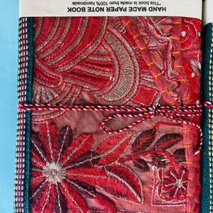 May include: A handmade paper notebook with a red embroidered cover featuring floral and paisley designs. The notebook is bound with a colourful cord and has a green border. The text on the cover reads "HAND MADE PAPER NOTE BOOK".