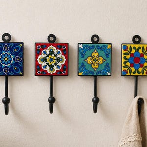 May include: Four decorative wall hooks with square, patterned ceramic tiles in blue, red, teal, and yellow. Each hook has a black metal frame and a curved hook. One hook holds a beige towel. The hooks are mounted on a white wall.