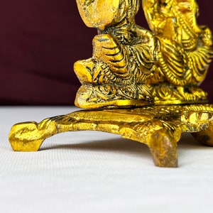 May include: A gold-colored metal decorative stand with two figures on top. The stand has a round top and three legs. The figures are intricately detailed and have a traditional design.
