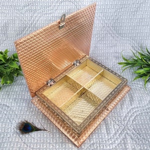 Può includere: Una scatola di legno color oro e rame con un coperchio incernierato e quattro scomparti. La scatola presenta un design decorativo all'esterno ed è aperta per rivelare gli scomparti.
