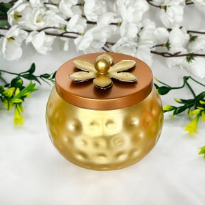 May include: A gold metal container with a hammered finish and a lid featuring a decorative flower design. The lid is copper coloured.