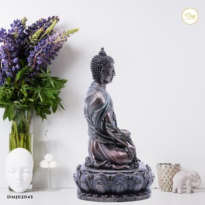 May include: A dark bronze Buddha statue sitting in meditation pose on a lotus base. The statue has a detailed face and intricate clothing. The base is decorated with a lotus flower design.