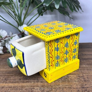 May include: A small yellow wooden box with a hand-painted design. The box has a drawer that is pulled out, revealing a white ceramic interior. The drawer has a floral design in blue, green, and yellow.