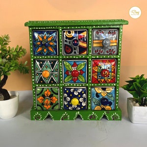 Puede incluir: Una caja de madera verde con doce cajones pequeños. Cada cajón está decorado con un diseño de azulejos de colores diferente con patrones geométricos y flores.