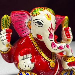 May include: A red, white and gold painted figurine of the Hindu deity Ganesha. The figurine is sitting with its trunk curled up and its hands in a gesture of blessing. The figurine is decorated with intricate details and colourful paint.
