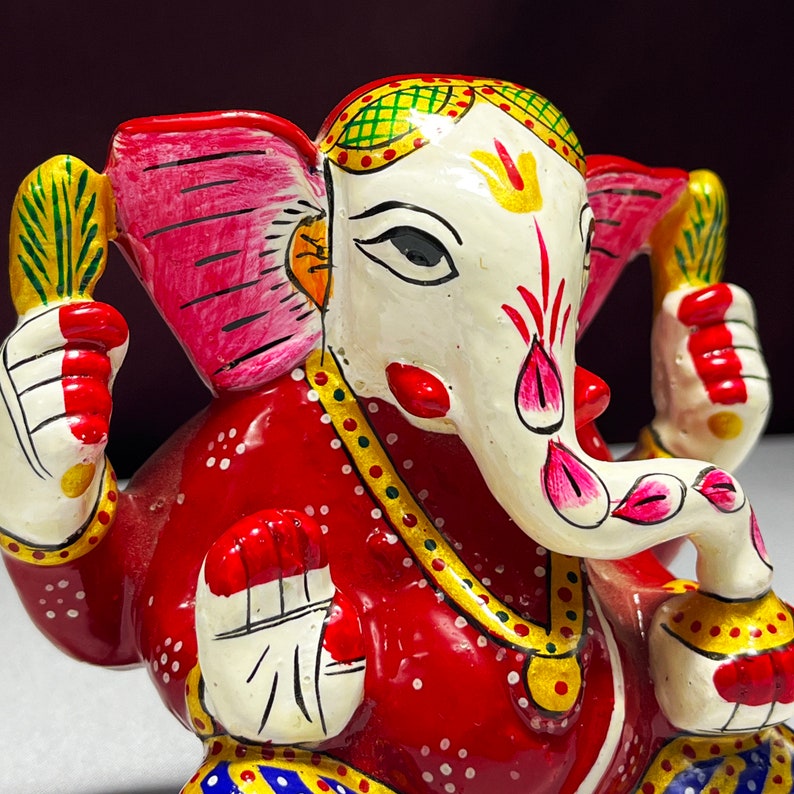 May include: A red, white and gold painted figurine of the Hindu deity Ganesha. The figurine is sitting with its trunk curled up and its hands in a gesture of blessing. The figurine is decorated with intricate details and colourful paint.