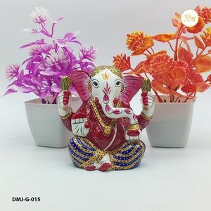 May include: A colourful metal statue of the Hindu deity Ganesha, sitting with his trunk raised and holding a sweet in his hand. The statue is painted in red, white, gold, and blue, and is decorated with intricate details. The statue is sitting on a white surface, with two potted plants in the background.