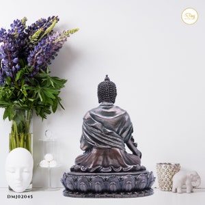 May include: A bronze Buddha statue sitting in meditation pose on a lotus base. The statue has a dark brown patina and is decorated with intricate details. The statue is facing away from the viewer.