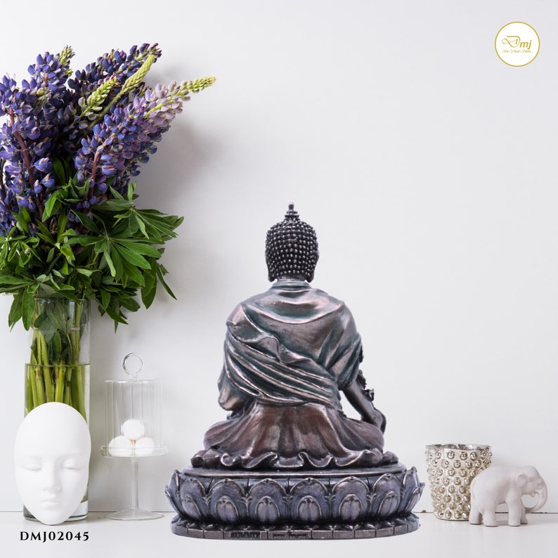 May include: A bronze Buddha statue sitting in meditation pose on a lotus base. The statue has a dark brown patina and is decorated with intricate details. The statue is facing away from the viewer.