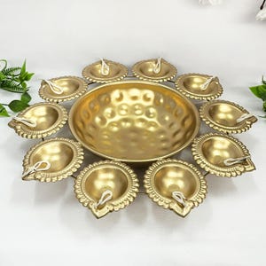 Op de afbeelding: Een gouden metalen diya met 10 individuele diyas die in een cirkel rond een grotere centrale diya zijn gerangschikt. De diyas zijn allemaal versierd met een parelrand.
