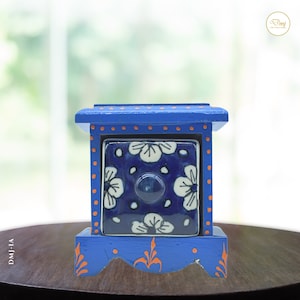 Op de afbeelding: Een kleine blauwe houten doos met een blauwe en witte keramische lade. De lade heeft een bloemenpatroon en een kleine blauwe knop. De doos is versierd met oranje stippen en bloemmotieven.