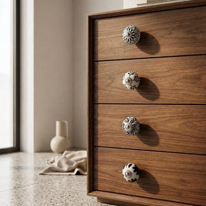 May include: A wooden chest of drawers with four drawers, each fitted with a decorative ceramic knob. The knobs display black and white floral designs. The chest is made of warm-toned wood.