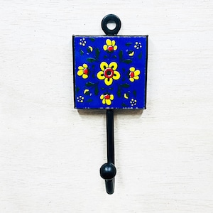 Könnte beinhalten: Ein dekorativer Wandhaken mit einer quadratischen, blauen Keramikfliese mit einem floralen Design in Gelb und Rot. Der Haken ist aus schwarzem Metall gefertigt und hat oben eine Schlaufe zum Aufhängen. Der Haken ist ca. 15 cm lang.