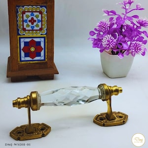 Puede incluir: Una manija de puerta de vidrio transparente con un diseño facetado y detalles en metal dorado. La manija está montada en dos placas de metal dorado con bordes decorativos. La manija está etiquetada como "DMJ-WSDH-01".