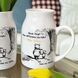 May include: Two white ceramic mugs with handles, each decorated with a black and white illustration of a bear and a smaller animal under a tree. The mugs have the text "Never forget the difference you make" and "Thank you Sophia". One mug holds yellow tulips.