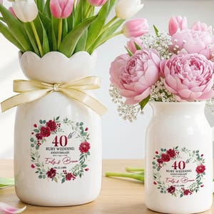 May include: Two white ceramic vases with floral arrangements. One vase has a scalloped top and a gold ribbon. Both vases feature a wreath design with text that reads "40th Ruby Wedding Anniversary". The vases are filled with pink and white tulips and pink peonies.