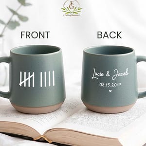 May include: Two sage green ceramic mugs with speckled textures and unglazed tan bases. The front mug features white tally marks, while the back mug displays the white text "Lucie & Jacob" above the date "08.15.2017" and a small heart icon.