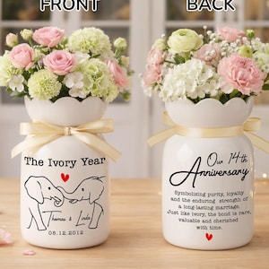May include: Two white ceramic vases with floral arrangements. The front vase has the text "The Ivory Year" and elephant illustrations. The back vase says "Our 14th Anniversary" with text about marriage. Both vases have cream-colored bows.
