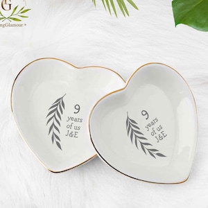 May include: Two heart-shaped, white ceramic dishes with gold rims. Each dish has a gray leaf design and the text "9 years of us J&E". The dishes are on a white, fluffy surface.