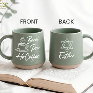 May include: Two green ceramic mugs with a tan base are displayed open on a book. The front mug has the text "Borei Pri HaCoffee" with a coffee cup graphic. The back mug has a Star of David and the name "Esther".