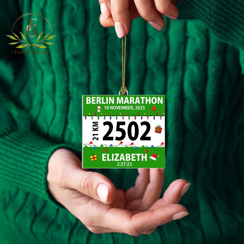 May include: A square Christmas ornament featuring a Berlin Marathon race bib design with the text BERLIN MARATHON, 10 NOVEMBER, 2025, 21 KM, 2502, ELIZABETH, and 2:27:23. The green and white design includes holiday lights, a snowman, and gifts.