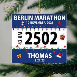 May include: A square Christmas ornament featuring a blue and white design with the text BERLIN MARATHON, 10 NOVEMBER, 2025, 42 KM, 2502, THOMAS, and 2:27:23. The ornament includes graphics of a snowman, Christmas tree, lights, and gifts.