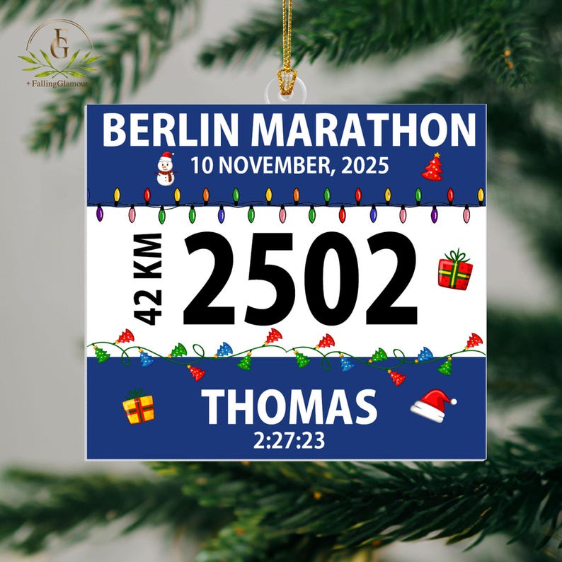 May include: A square Christmas ornament featuring a blue and white design with the text BERLIN MARATHON, 10 NOVEMBER, 2025, 42 KM, 2502, THOMAS, and 2:27:23. The ornament includes graphics of a snowman, Christmas tree, lights, and gifts.