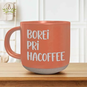 May include: A coral-coloured ceramic mug with a grey base and a curved handle. The mug features the white text "BOREI PRI HACOFFEE". The mug is sitting on a wooden surface.