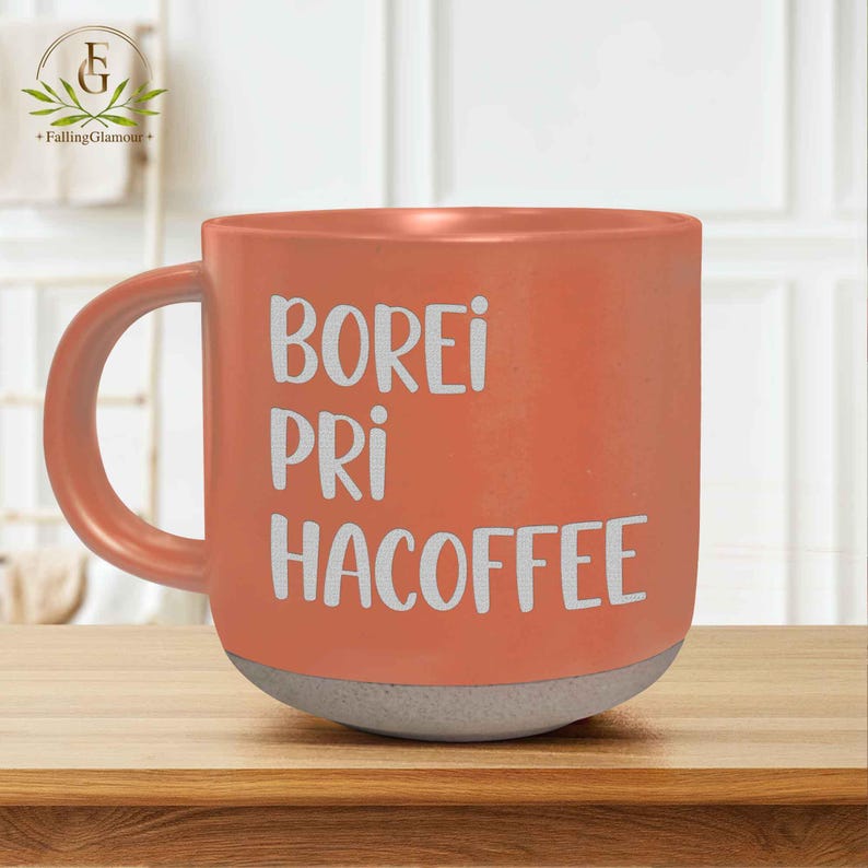 May include: A coral-coloured ceramic mug with a grey base and a curved handle. The mug features the white text "BOREI PRI HACOFFEE". The mug is sitting on a wooden surface.