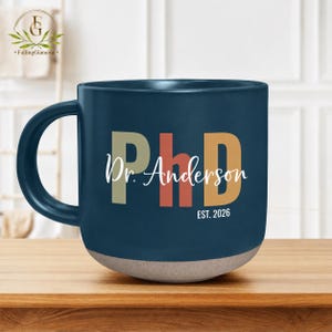 May include: A navy blue ceramic mug with a handle, featuring the text "PhD Dr. Anderson EST. 2026" in white, beige, orange, and brown. The bottom of the mug is a light beige color.