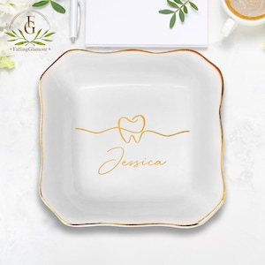 May include: White ceramic trinket tray with a gold rim and a gold outline of a tooth with a heart in the center. The name "Jessica" is written in gold script below the tooth.